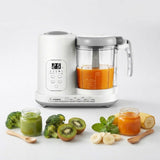 A white and gray baby food maker with a clear container filled with orange puree, shown on a light gray background with baby food jars, broccoli, kiwi, and banana slices arranged in front.